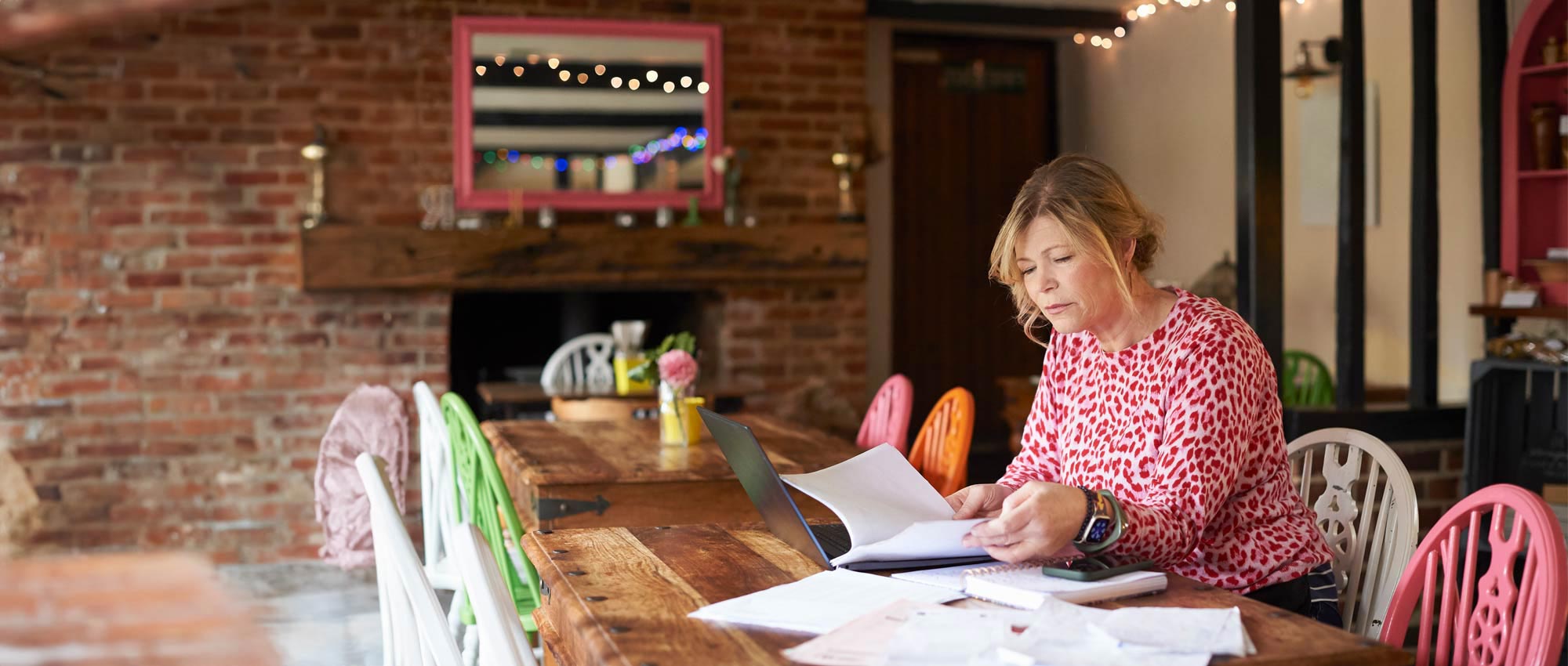 A woman is sitting reading some documents and looking at a laptop computer